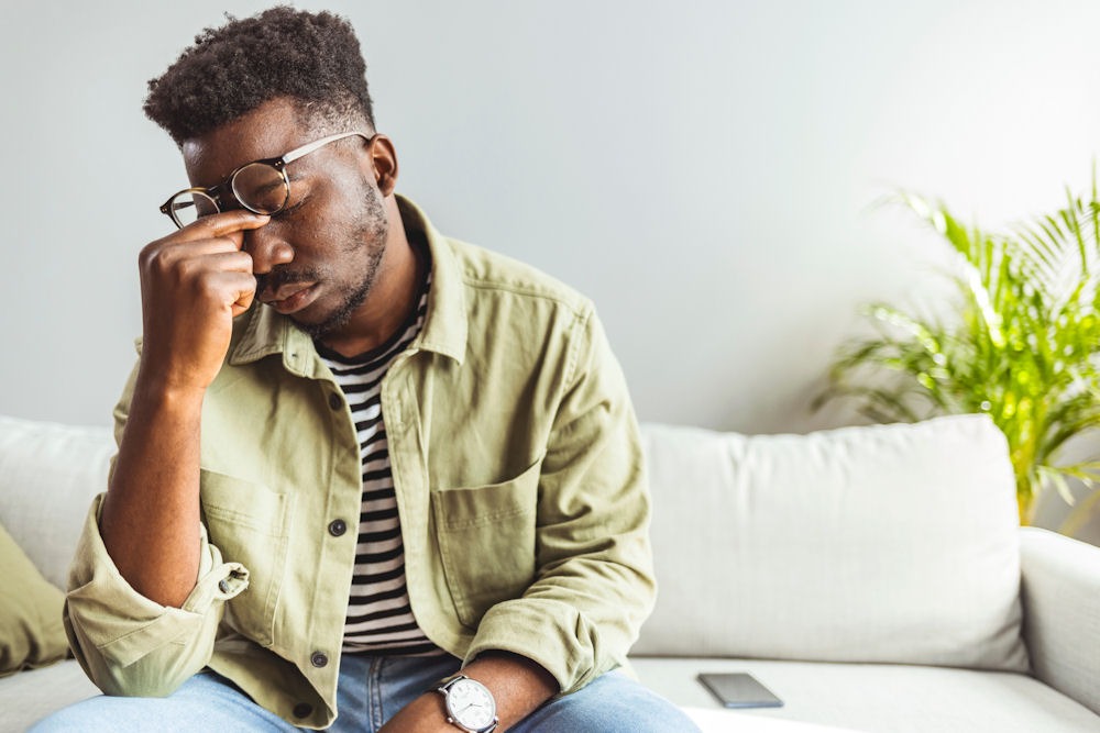 man sitting on white couch pinching the bridge of his nose in frusteration