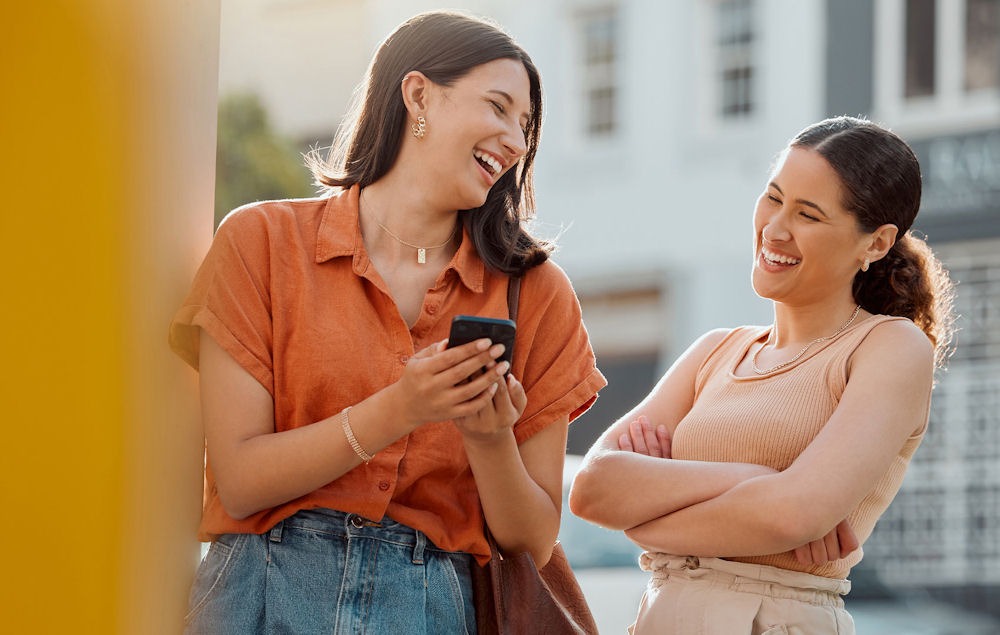 two women outside laughing while looking at one's phone