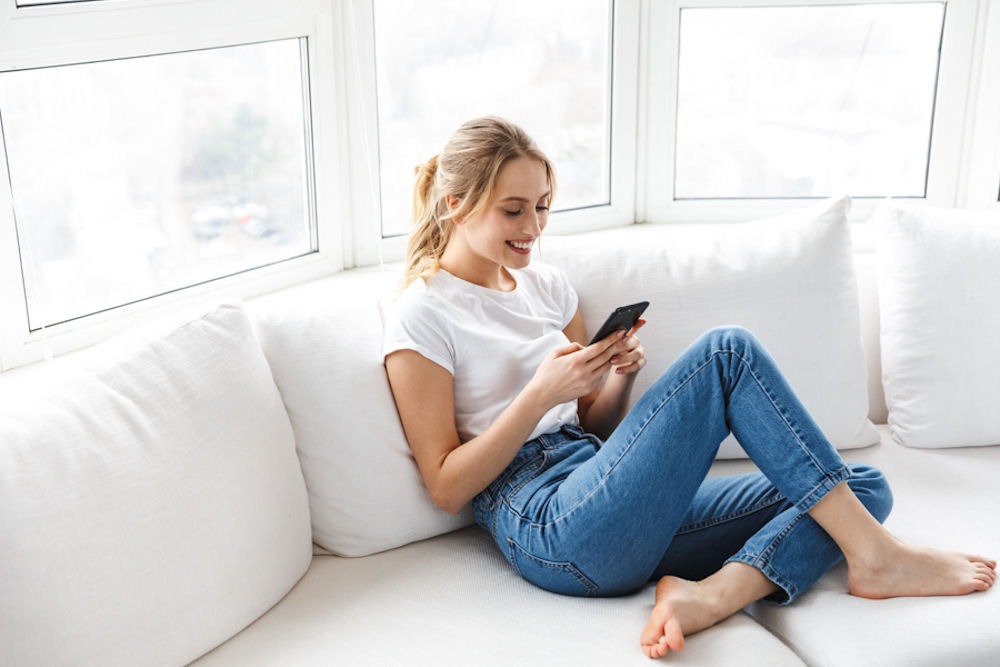 happy woman sitting on white couch looking at her phone